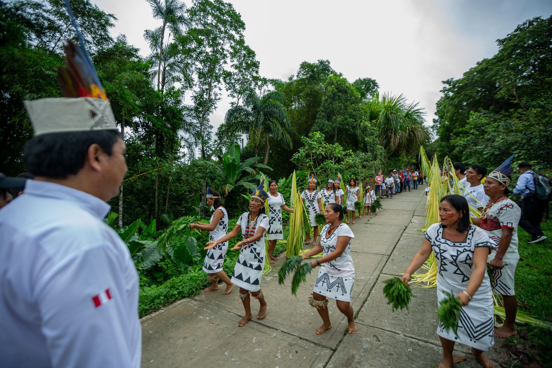 Members of an indigenous community in traditional clothing with white and black geometric motifs walk along a concrete path through dense tropical rainforest, holding green branches, while a man in white and blue uniform observes from the foreground.