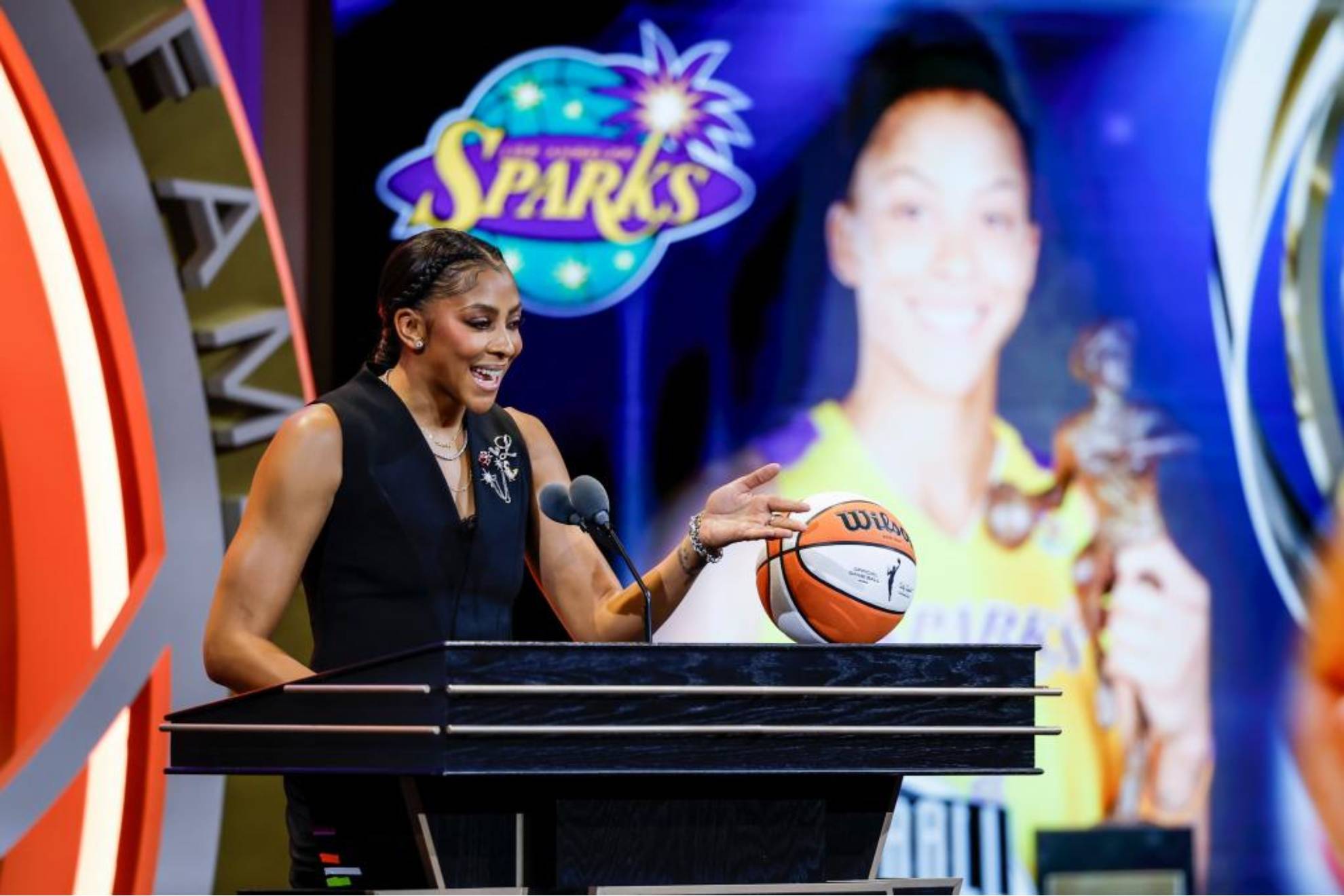 Una mujer en ropa negra de gala sostiene un balón de baloncesto mientras habla frente a un podio con micrófono, con el logo de los Sparks y una pantalla de video de fondo.