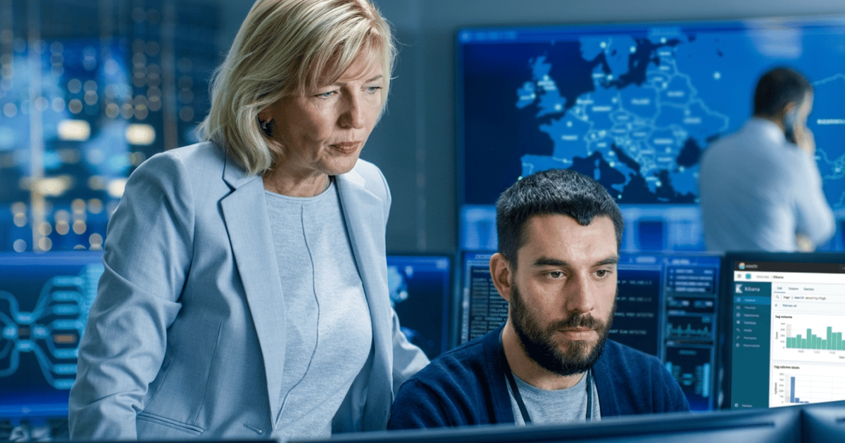 A woman and a man work together at computer stations in a high-tech control room filled with digital displays showing maps and data analytics.