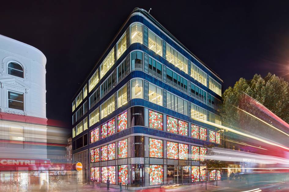 A modern multi-story building with blue and glass facade illuminated at night, with colourful floral-patterned windows on lower floors and bright interior lighting visible throughout.