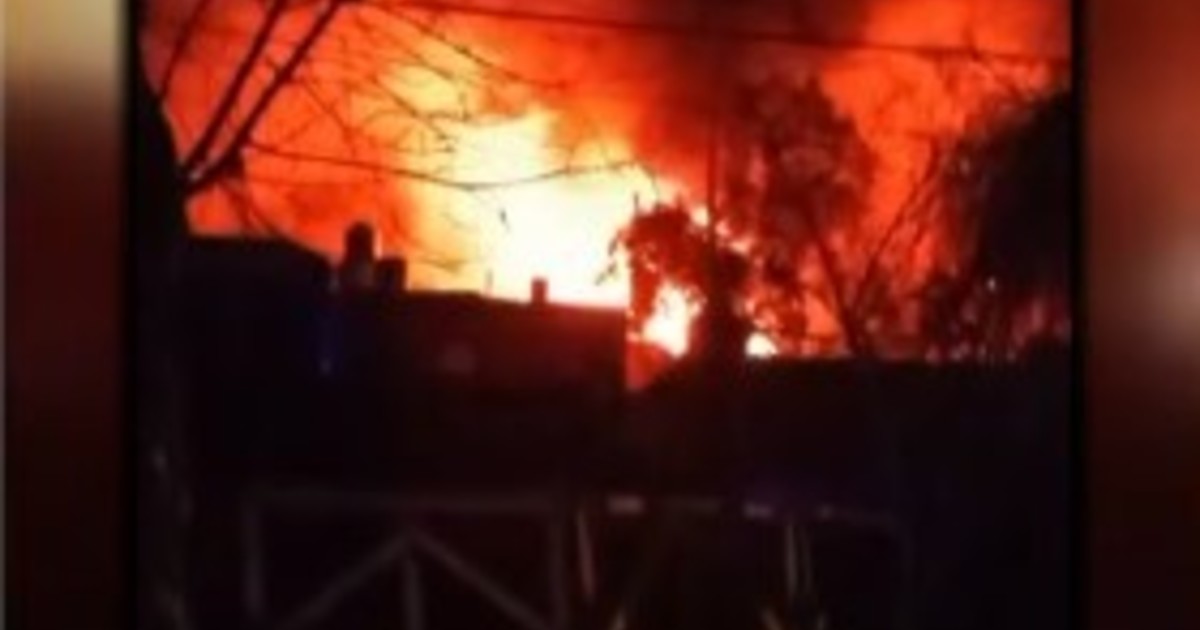 Un gran incendio ilumina la noche con llamas rojas y naranjas en lo que parece ser instalaciones industriales, con siluetas de estructuras contra el fuego intenso.