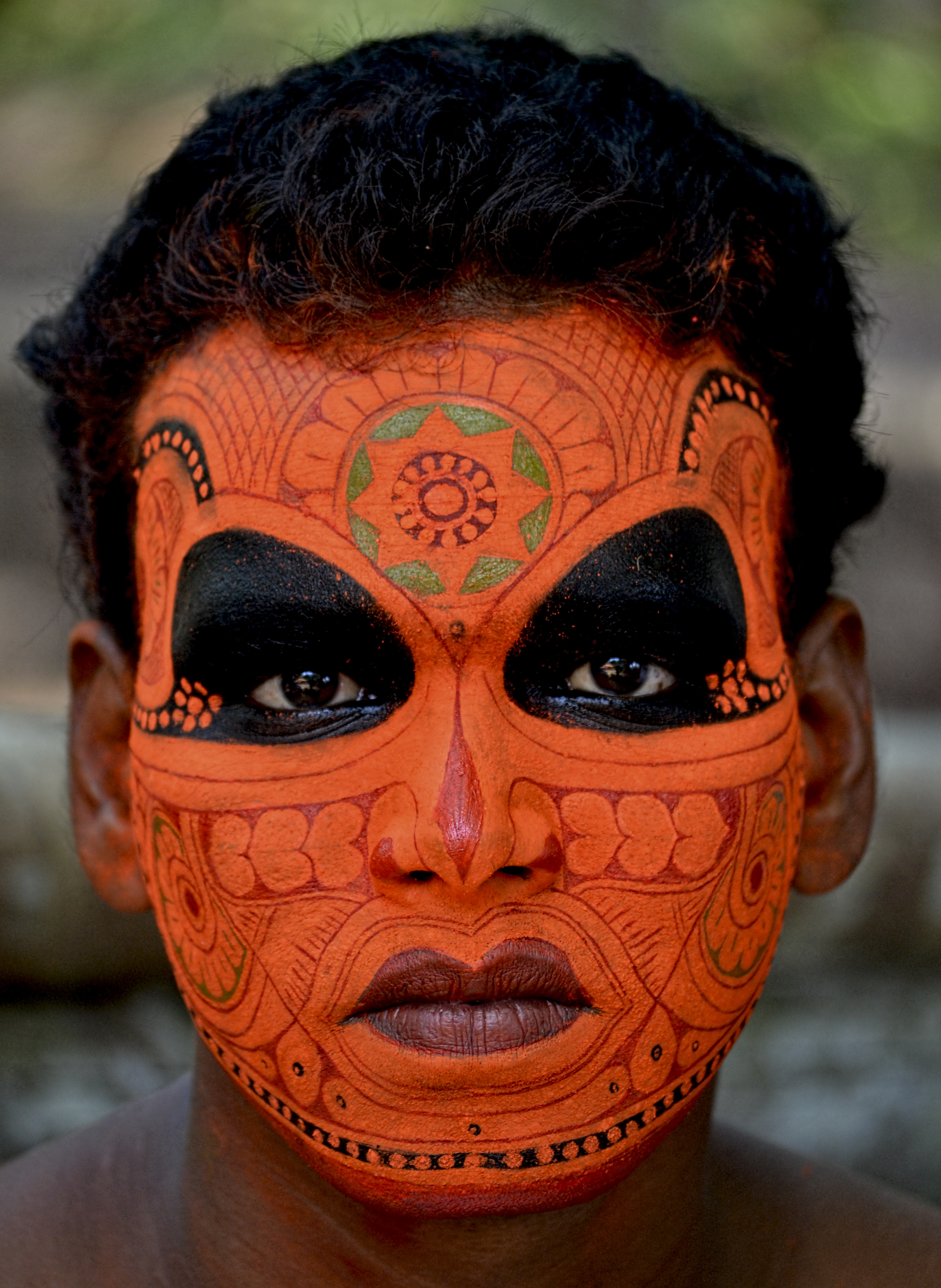 A person with traditional facial body art painted in orange and black, featuring intricate geometric patterns and designs covering the entire face.