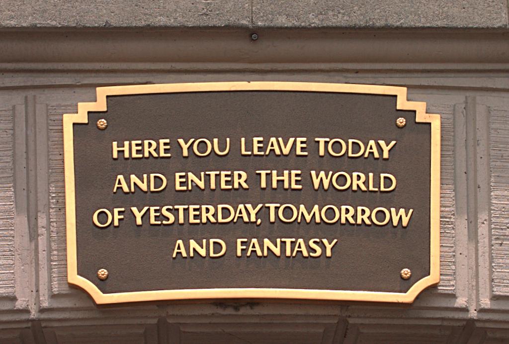 A bronze plaque with gold lettering mounted on a wooden panel reads "Here you leave today and enter the world of yesterday, tomorrow and fantasy."