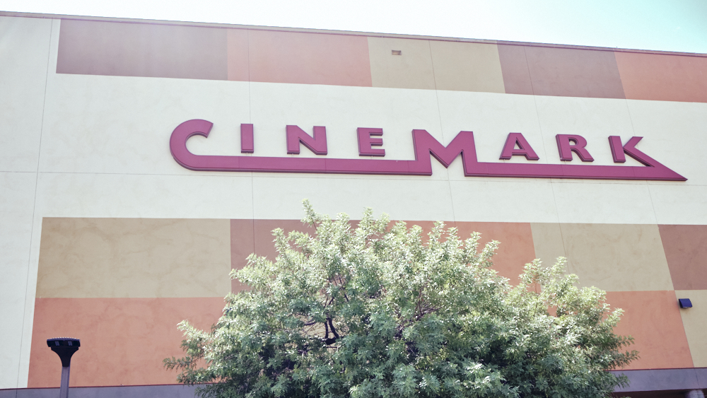 Fachada de un cine Cinemark con el letrero de la marca en letras púrpuras sobre una pared blanca, con un árbol verde en el primer plano.
