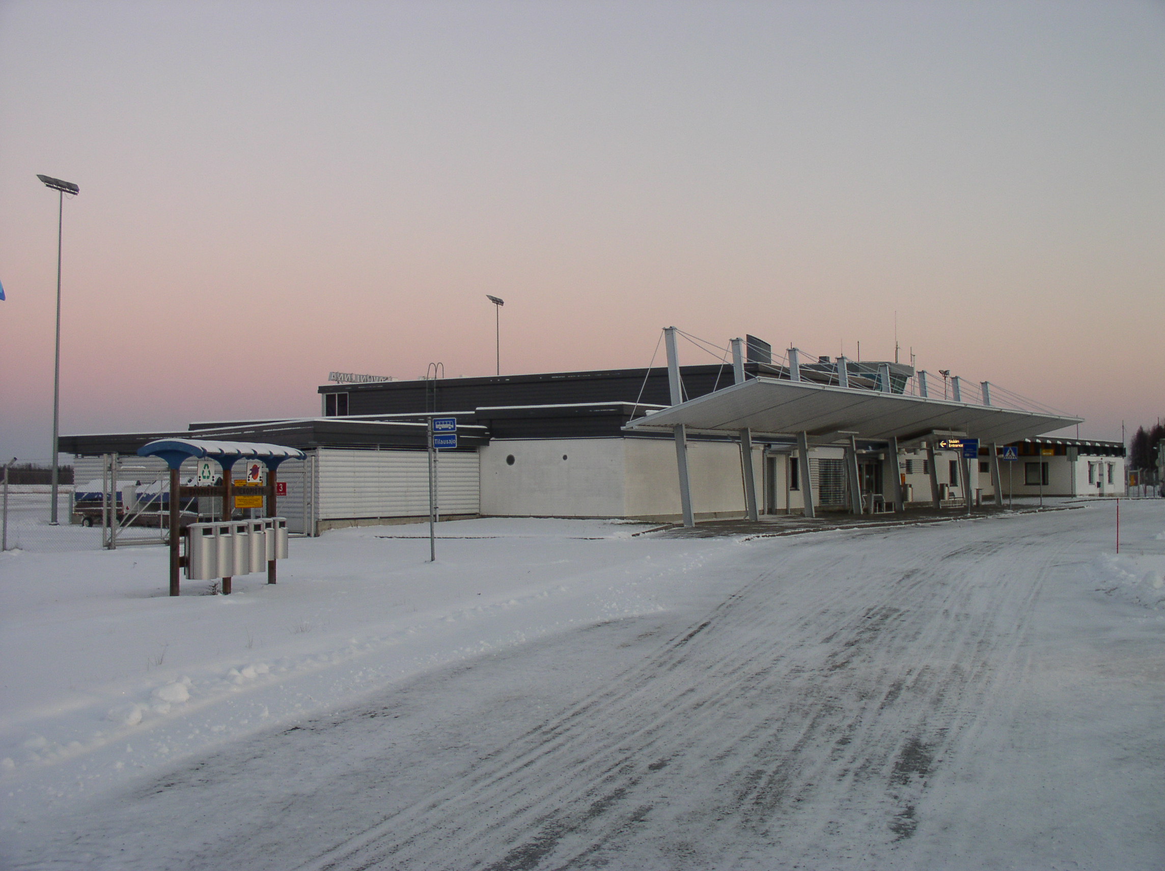 A modern commercial or industrial building with white walls and dark roof structures sits in a snowy landscape at dusk, with street lamps and minimal vegetation visible in the background.