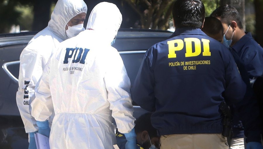 Varios investigadores de la PDI de Chile, algunos con trajes de protección blanca y otros con chalecos azul marino de la Policía de Investigaciones, trabajan en la escena de un crimen cerca de un vehículo.