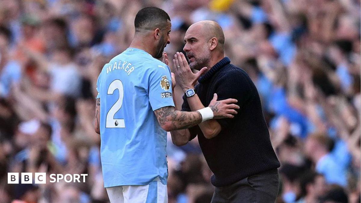 A manager in a black hoodie speaks with a Manchester City footballer wearing a light blue shirt with the number 2, during a match in front of spectators.