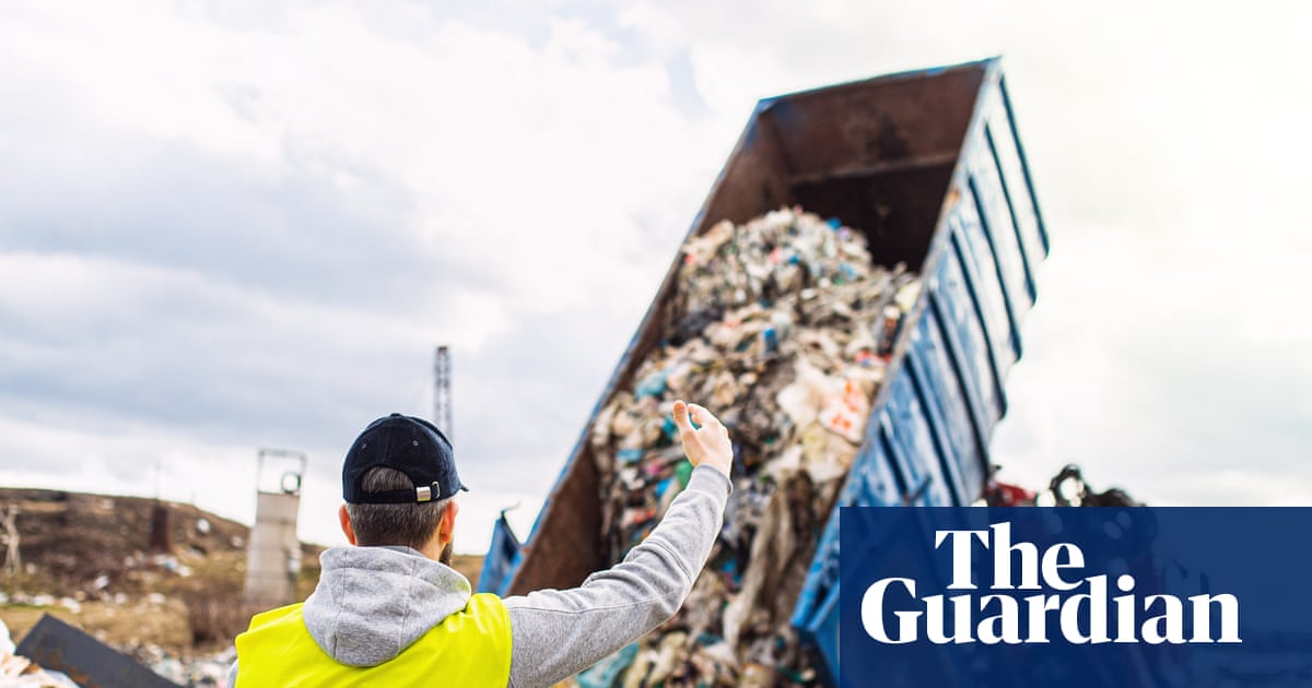 A worker in a yellow safety vest and black cap gestures toward a large blue dump truck tilted upward, discharging a massive load of mixed waste and recyclable materials.