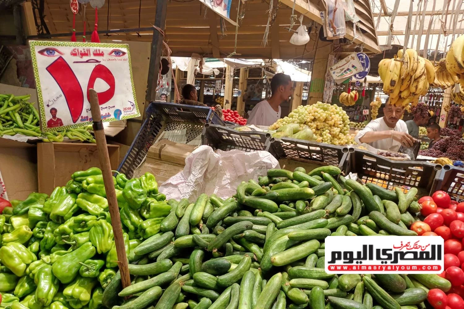 Un puesto de verduras en un mercado muestra abundantes pepinos, pimientos verdes, tomates rojos y otras frutas y verduras frescas con vendedores detrás del mostrador.