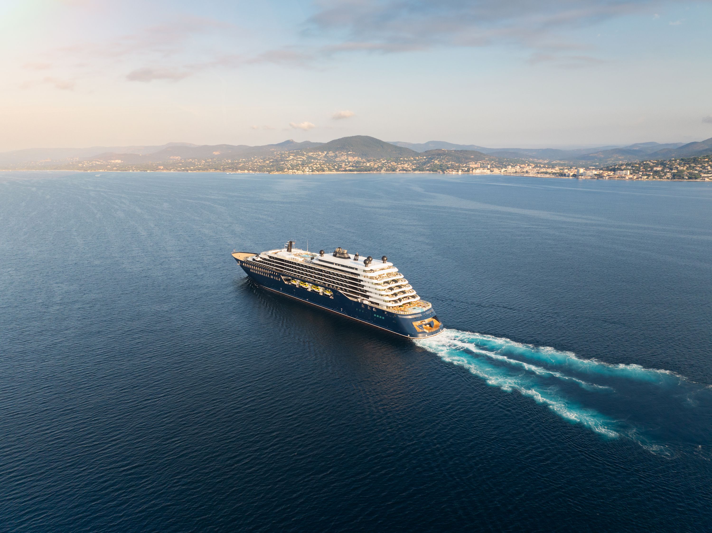 Un gran crucero de lujo blanco y azul navega en aguas del Mediterráneo con una costa montañosa y una ciudad costera de fondo.