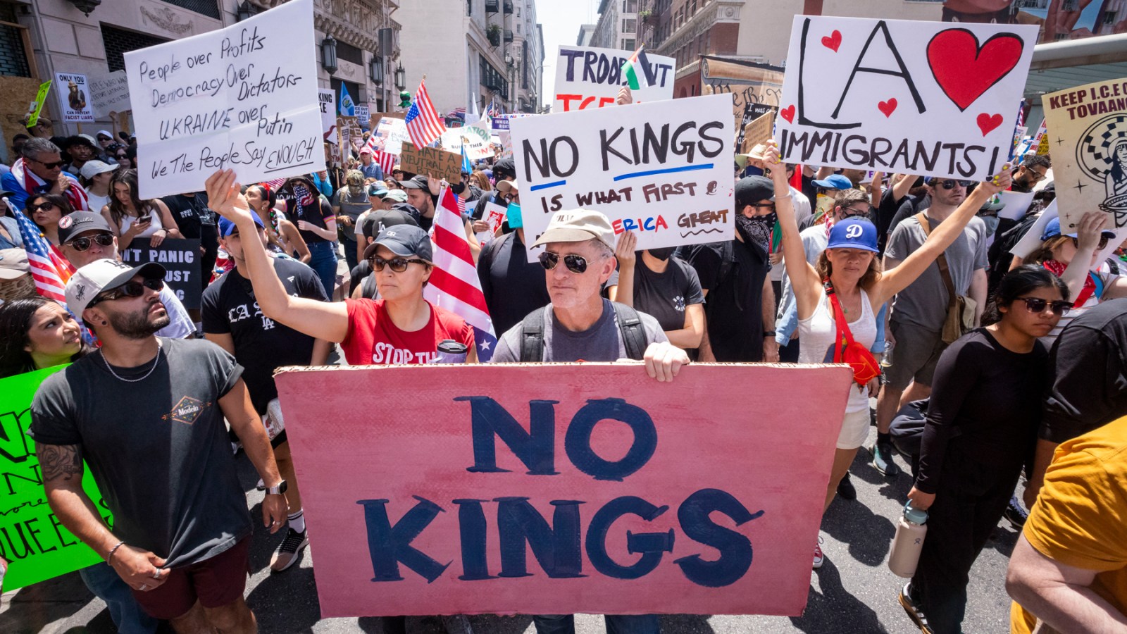 A large outdoor crowd of protesters holding signs and American flags on a city street lined with tall buildings.