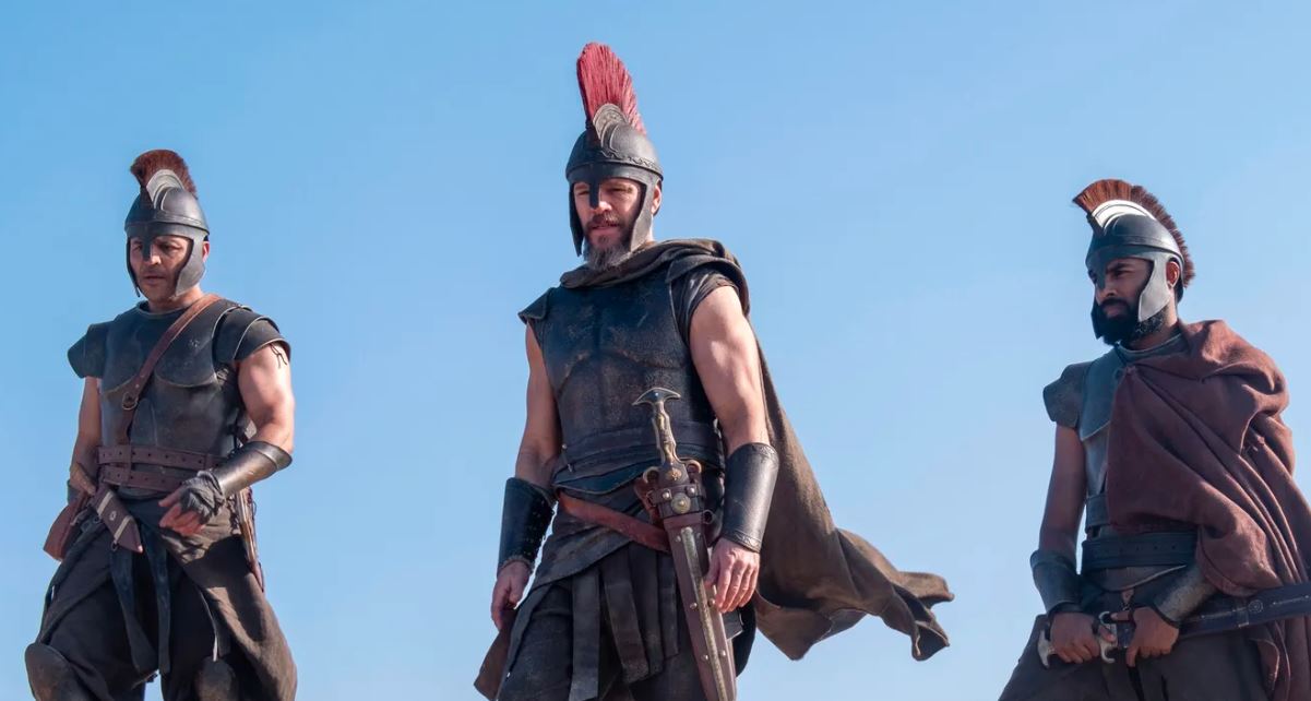 Three men dressed as ancient Greek warriors stand against a clear blue sky, wearing bronze helmets with red plumes, dark leather armor, and holding swords.