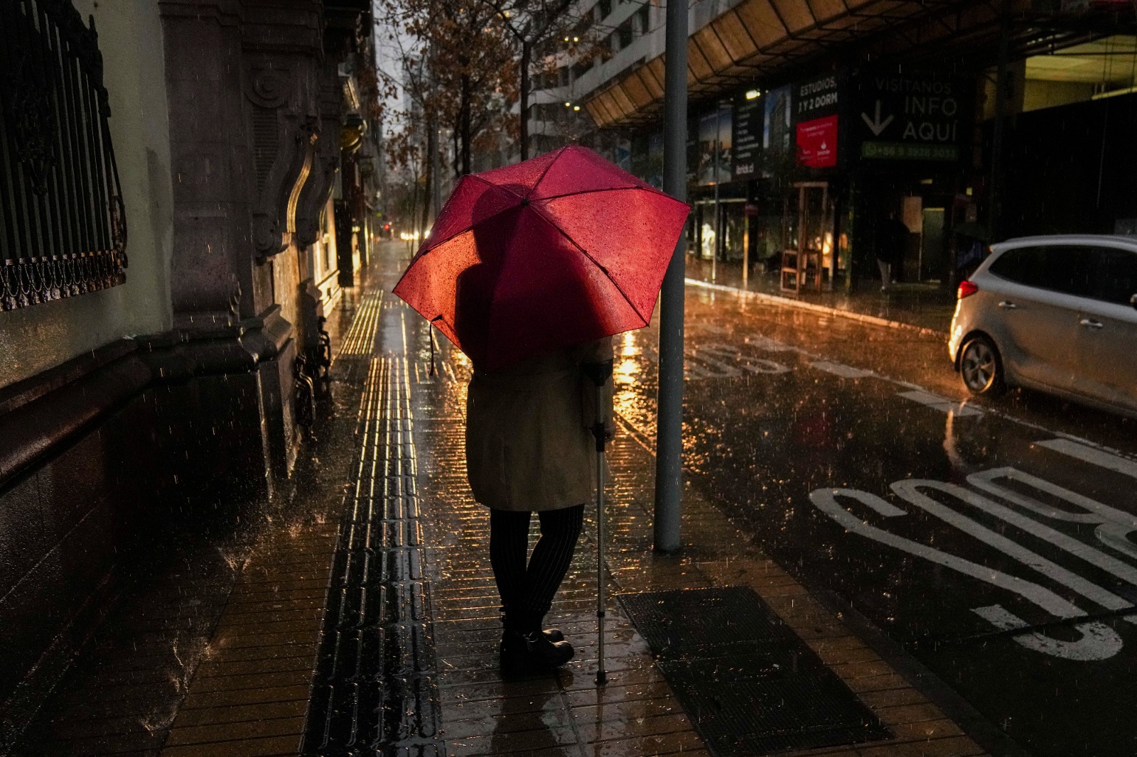 A person holding a red umbrella stands alone on a wet street during heavy rain, with a crosswalk and parked white car visible in the background.