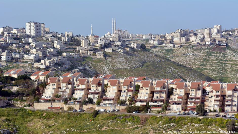Multi-storey residential houses with reddish-brown roofs built on a hillside, with green rocky terrain in the background and a larger urban settlement visible on higher hills.