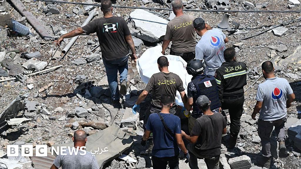 A group of people in rescue uniforms and civilians work among rubble and ruins of destroyed buildings, moving what appears to be a body covered with a white sheet.