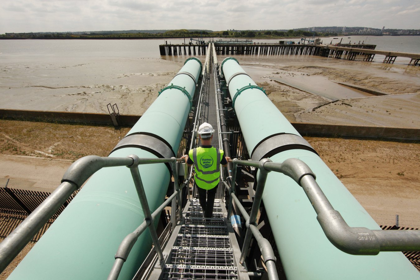 A worker in fluorescent yellow safety vest walks along a platform between two large pale green pipes at an industrial facility alongside an estuary, with damaged docks visible in brown water in the background.
