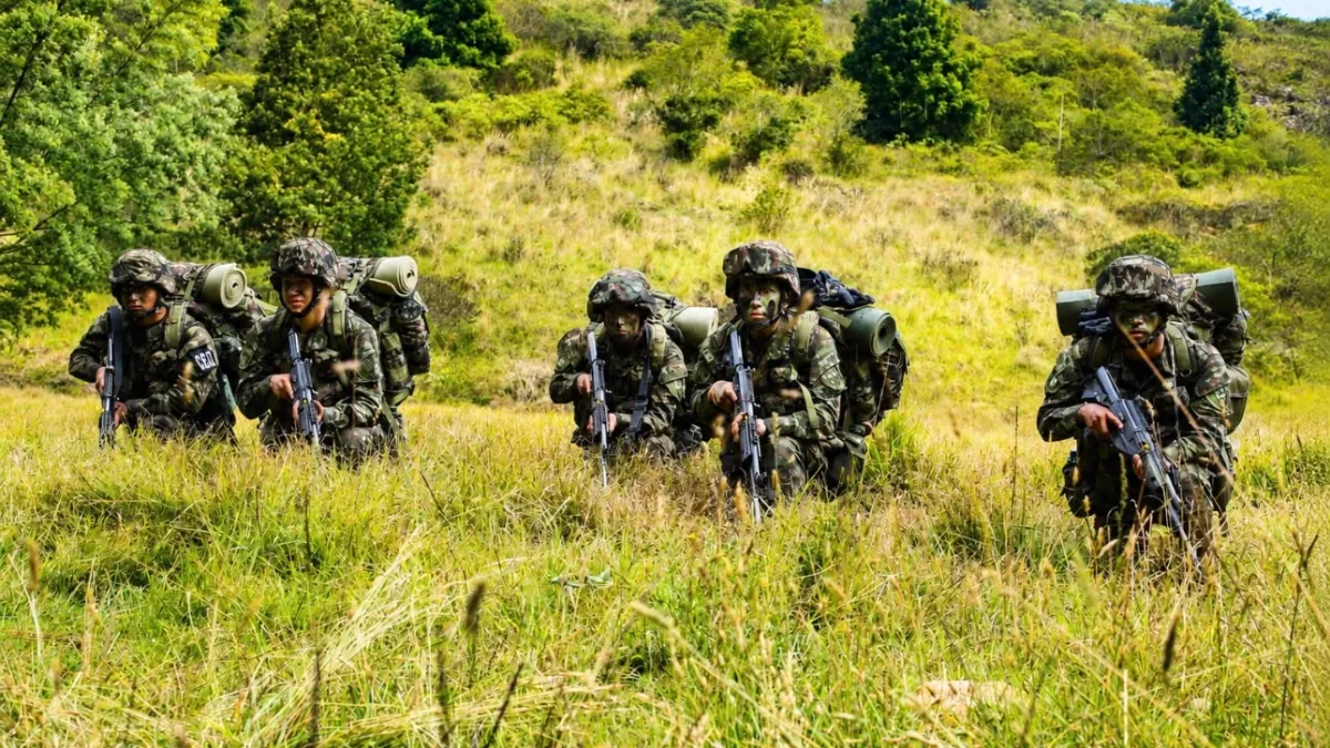 Un grupo de soldados armados con equipamiento militar avanza a través de un terreno cubierto de hierba verde con árboles y vegetación densa al fondo.