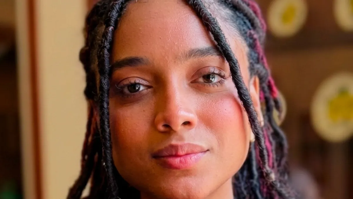 Portrait of a woman with black braids and pink details, looking directly at the camera with a serious expression indoors.