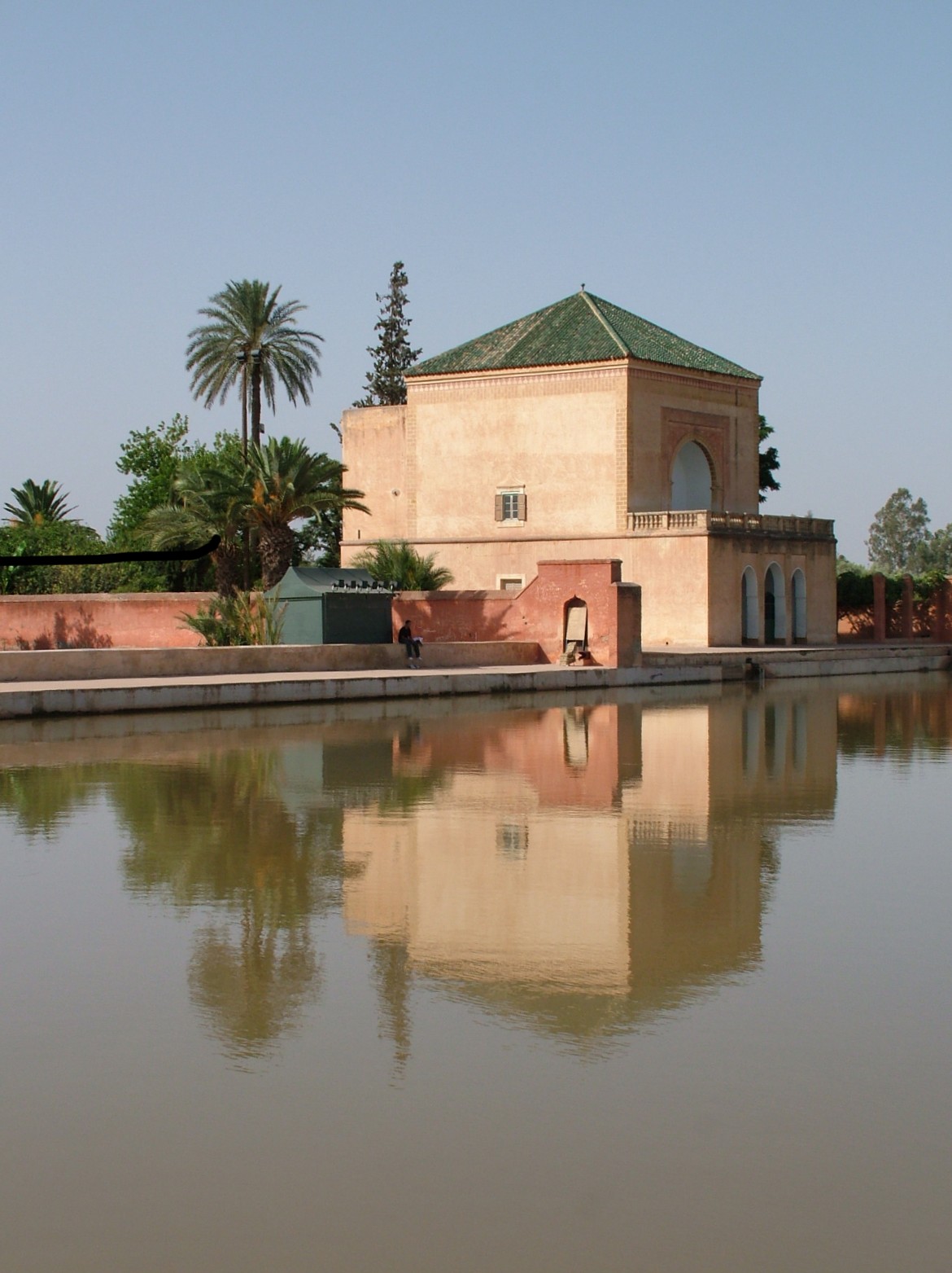 A pink-terracotta building with a green pyramidal roof sits beside a still body of water, surrounded by palm trees and manicured gardens in what appears to be a Moroccan palace or garden setting.