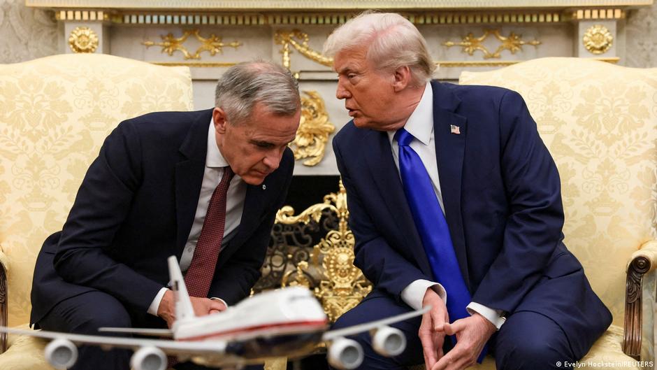 Two men in dark suits sitting in light-coloured upholstered armchairs, conversing in an ornately decorated room with gold details and classical decorations.