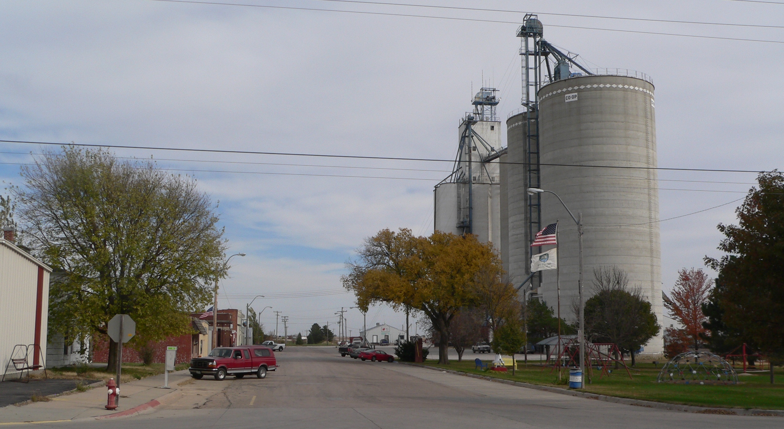 A wide main street in a small rural town with grain silos and industrial buildings visible in the background, trees showing fall foliage on both sides, and a few parked vehicles.