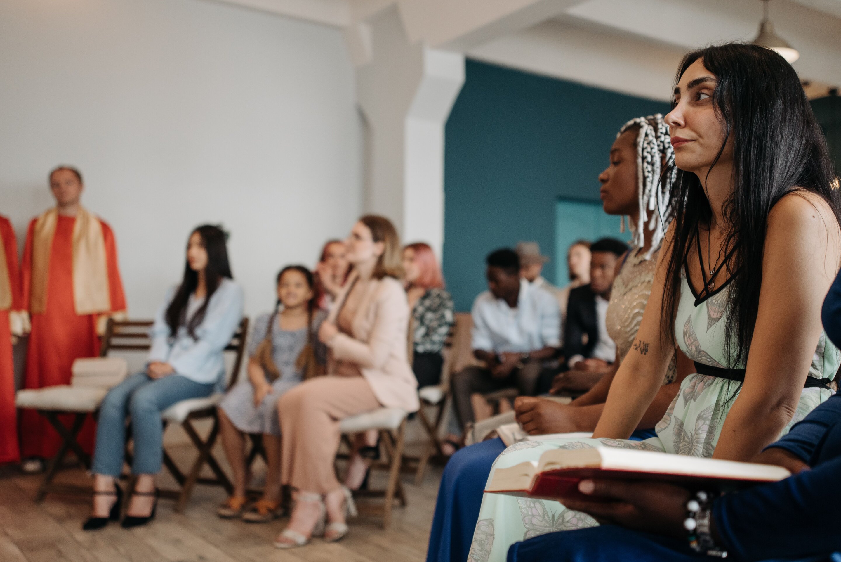 Un grupo diverso de personas se reúne en un círculo en una sala con paredes blancas y un panel azul, con algunos sosteniendo libros religiosos mientras escuchan a una líder de pie.