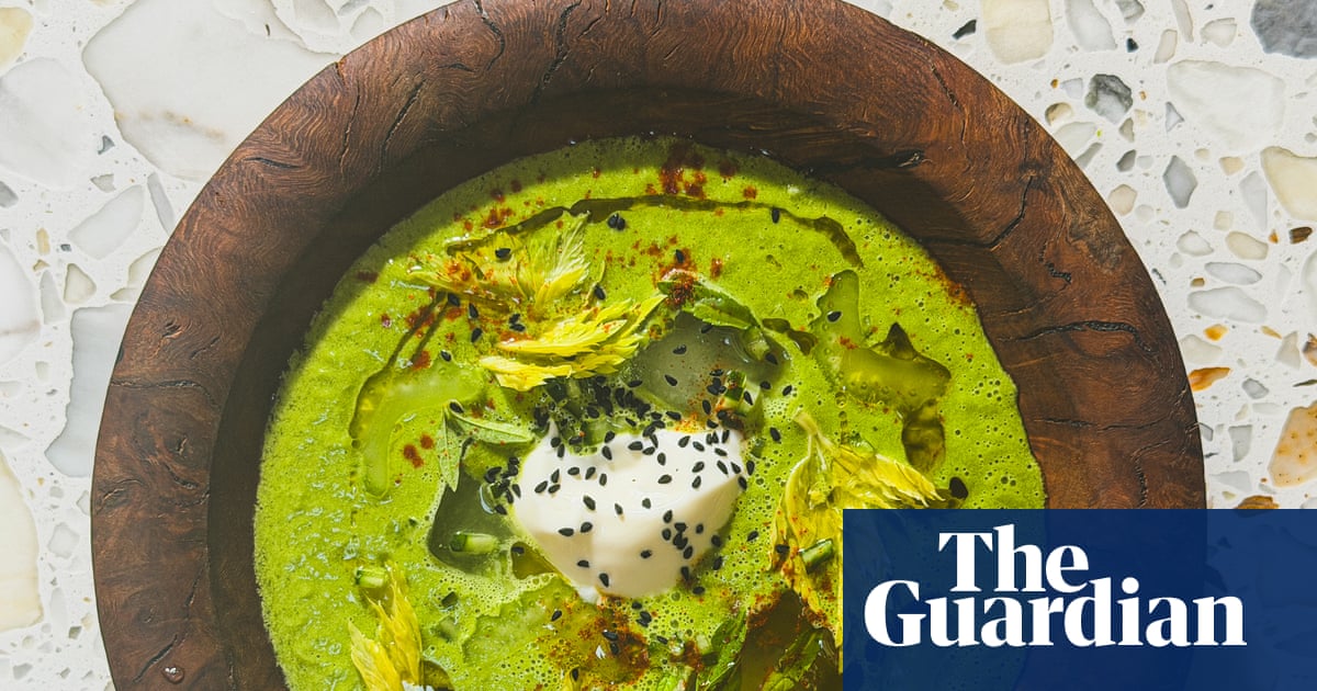 A bowl of green gazpacho with sour cream, black seeds and edible yellow flowers in a wooden container on a textured white mosaic surface.