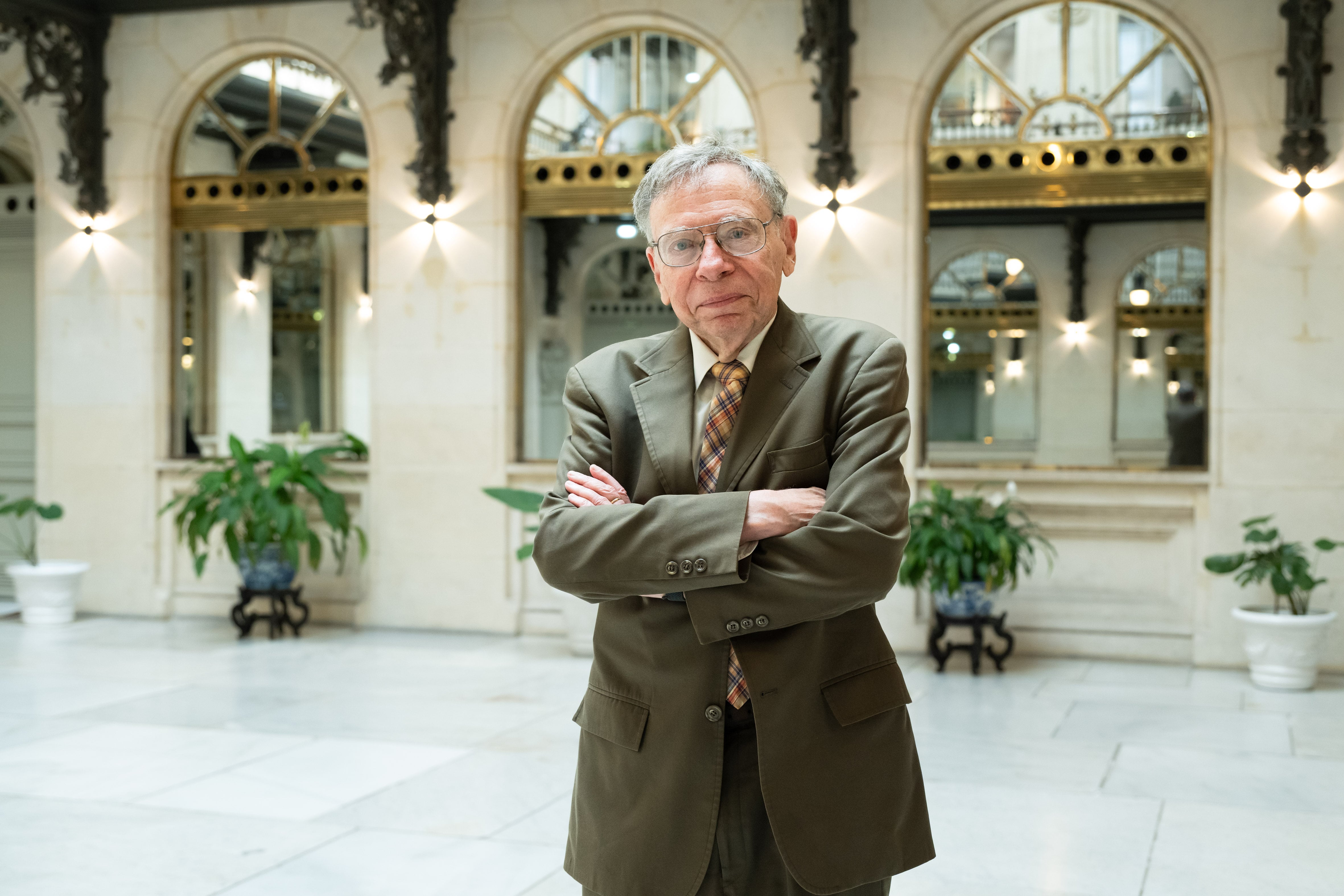 An older man with gray hair and glasses wearing a brown suit and plaid tie stands with crossed arms in an elegant corridor with arched windows and green plants.