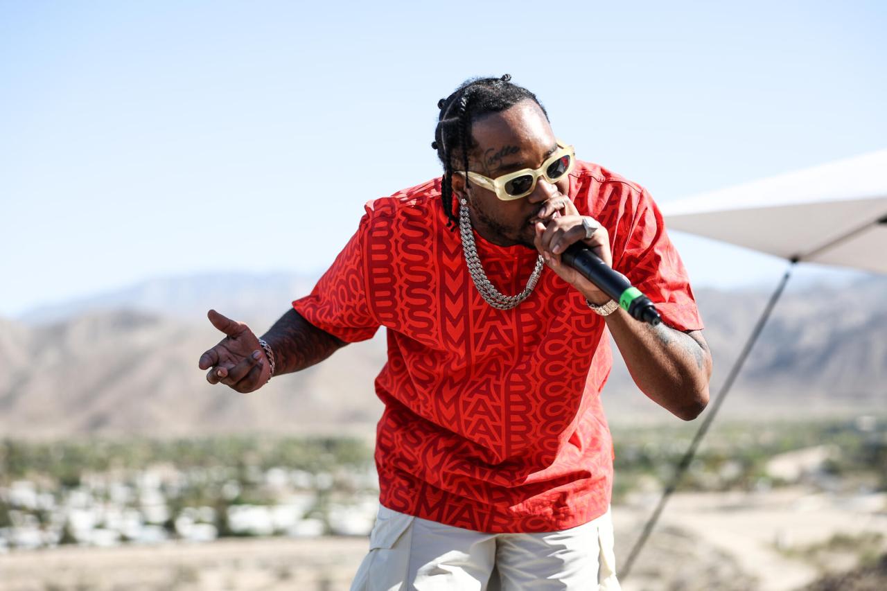 A man in a bright red patterned shirt and cream-colored sunglasses holds a microphone while performing, with a scenic landscape of white buildings and mountains visible in the background.