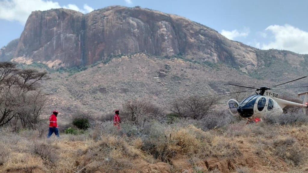 A white and blue helicopter with registration number 5Y-BUF sits on arid terrain with rescue workers in red vests nearby, with a distinctive flat-topped rocky mountain visible in the background.