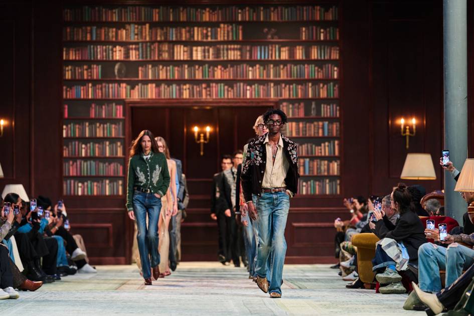 Modelos desfilan sobre una pasarela de moda en una biblioteca de ficción, con paredes llenas de libros de colores variados de fondo.