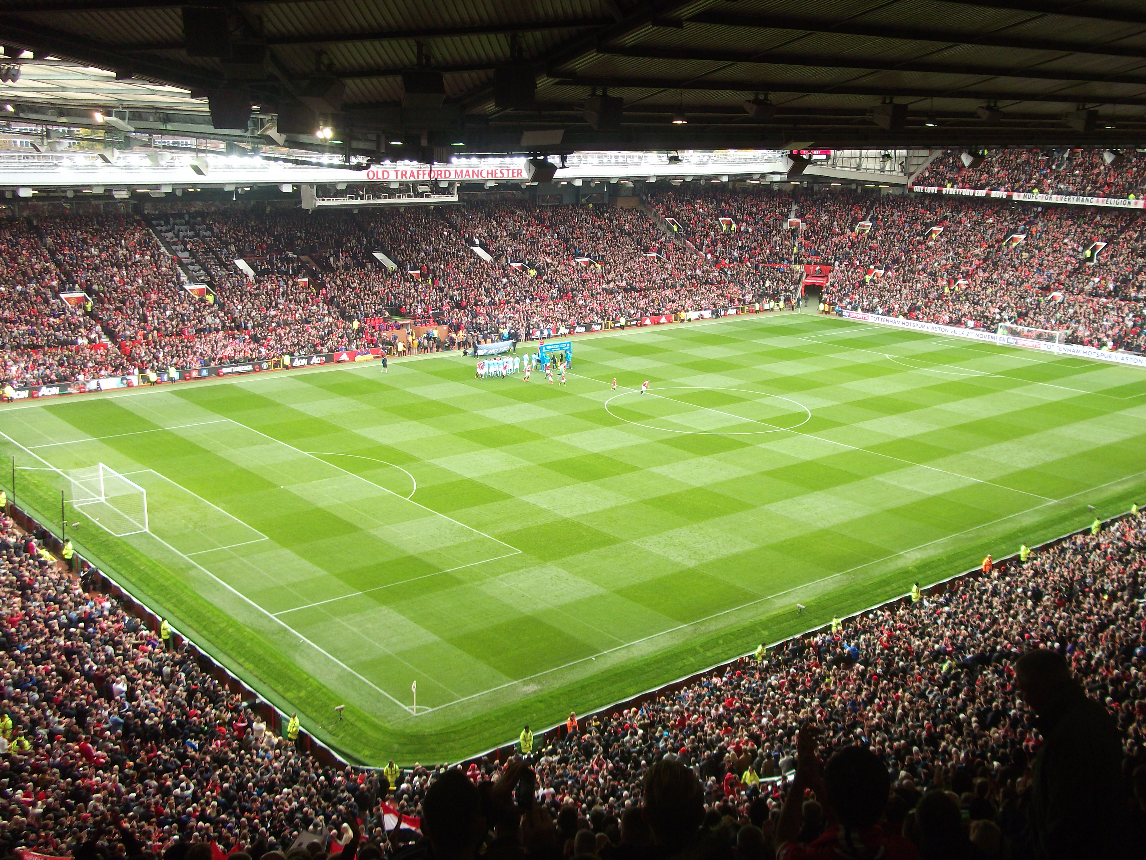Una vista aérea del Old Trafford, el estadio del Manchester United, lleno de aficionados durante un partido de fútbol, con el terreno de juego perfectamente delineado en verde.