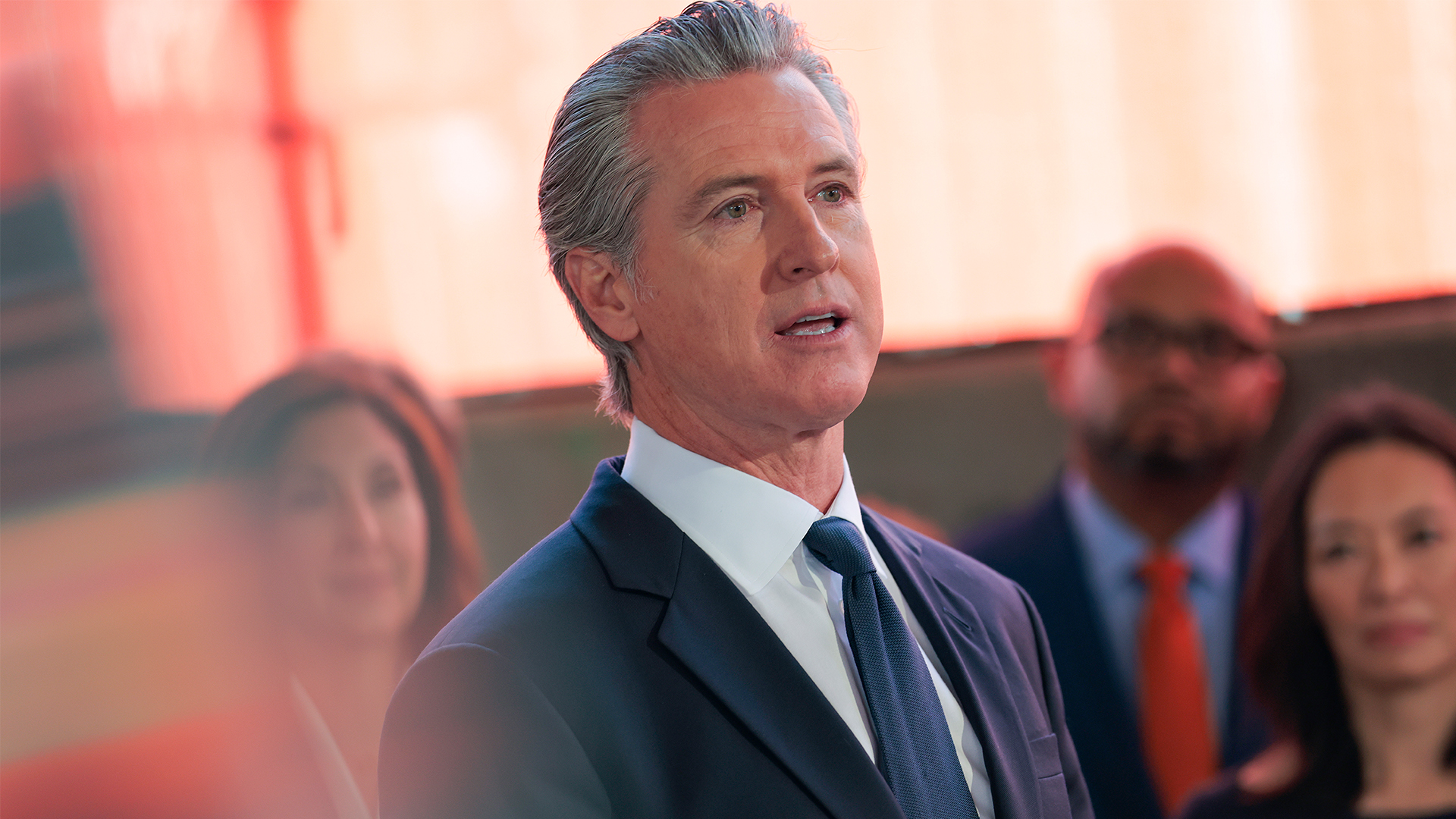 A man in a dark blue suit and tie stands in front of a blurred background with a reddish-orange hue, with other people visible out of focus behind him.