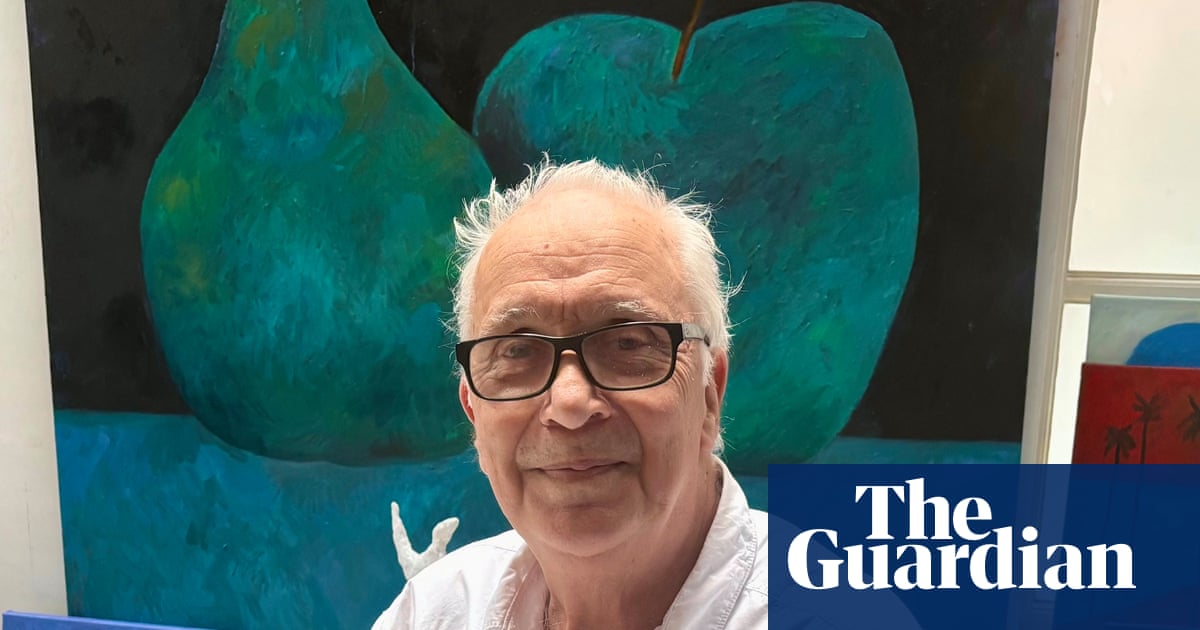 Un hombre mayor con gafas y cabello blanco, vistiendo una camisa blanca, posa frente a una obra de arte abstracta que muestra formas en tonos de azul y verde oscuro.