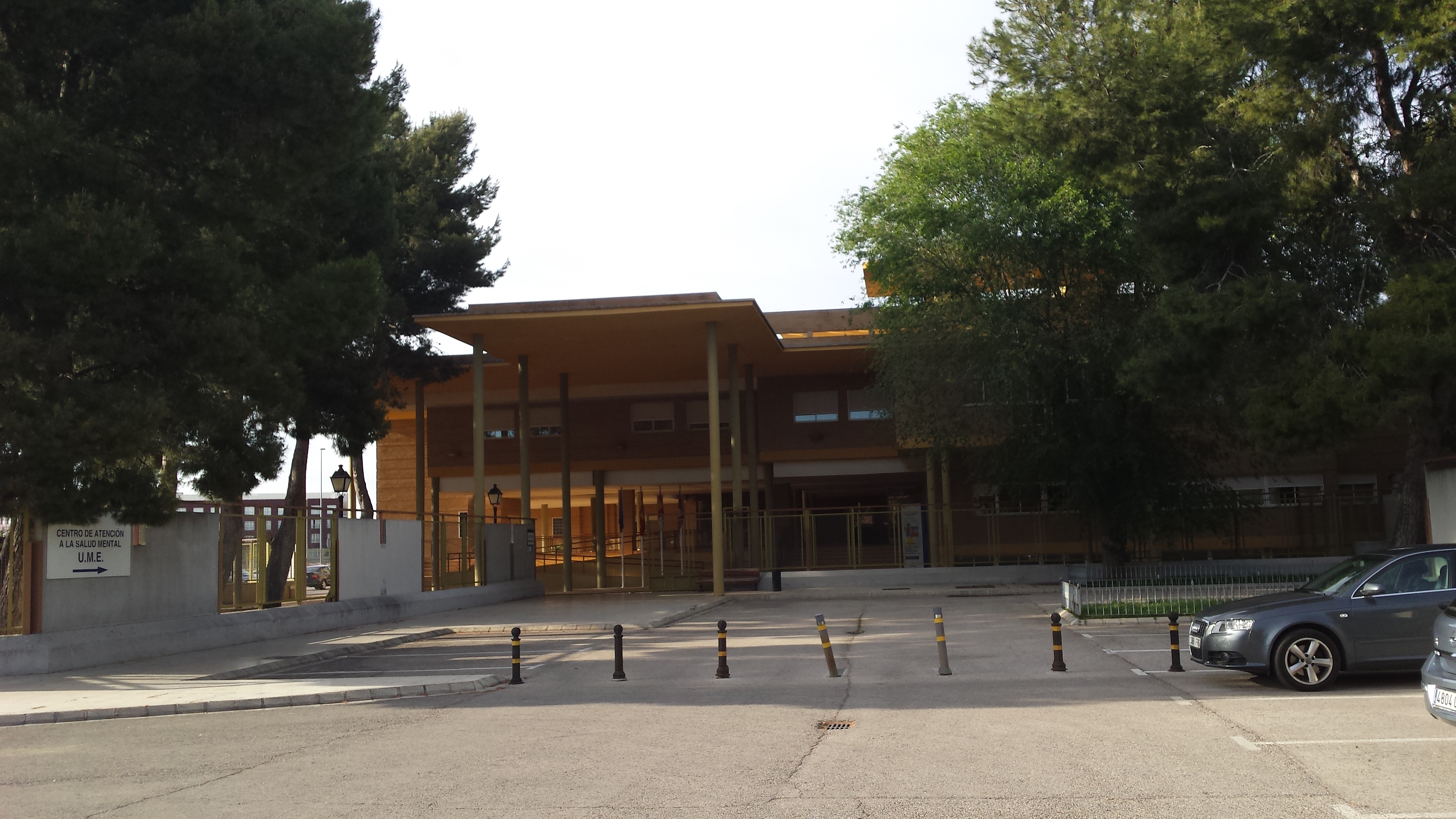 Exterior view of a modern school building with wooden cladding, concrete pillars, and large windows, surrounded by mature trees, with a courtyard area and parked cars visible in front.