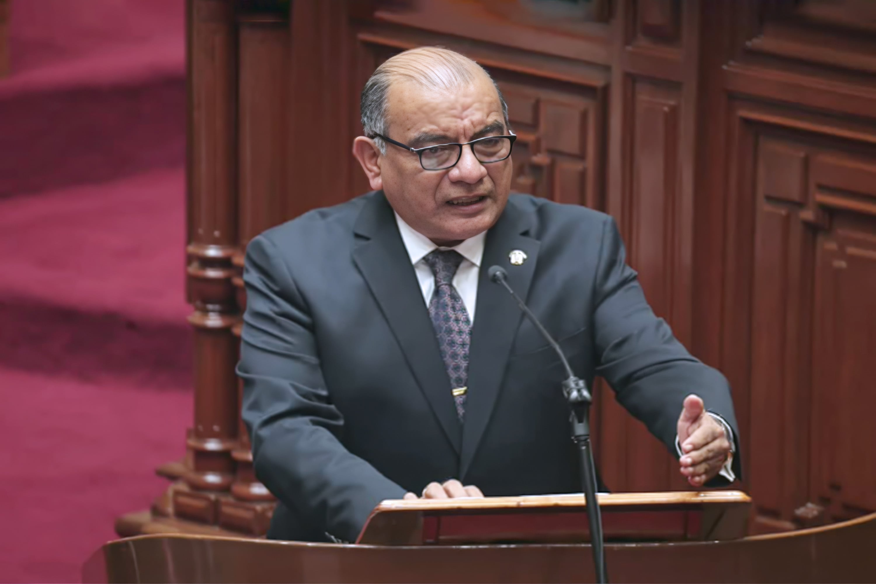 Un hombre de mediana edad con lentes, vestido con traje azul oscuro y corbata, habla desde un podio en lo que parece ser una sesión parlamentaria, con sus manos gesticulando mientras se dirige al micrófono.
