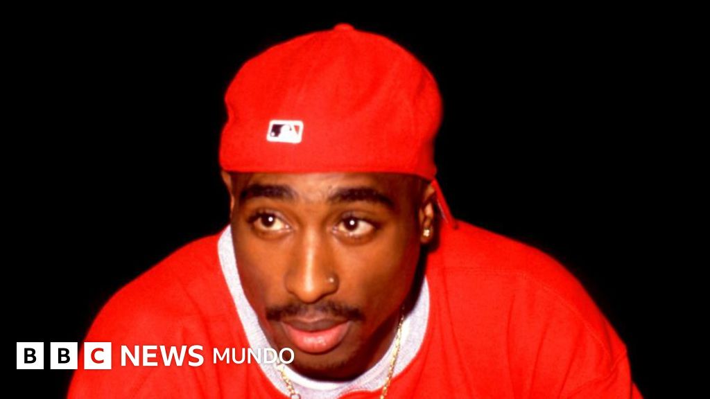 Portrait of a young man facing forward against a black background, wearing a red baseball cap with a Major League Baseball logo and a red sweatshirt with a white collar.