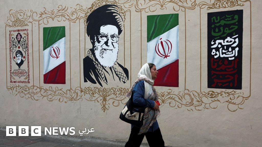 A woman walks past a mural on a wall depicting the portrait of an Iranian religious leader in black and white, flanked by Iranian flags and text in Persian.