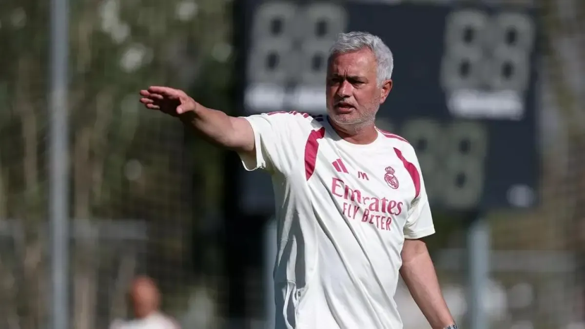 A middle-aged man with grey hair wearing a white and red Real Madrid shirt gestures with his arm extended during what appears to be a training session or work session on the pitch.