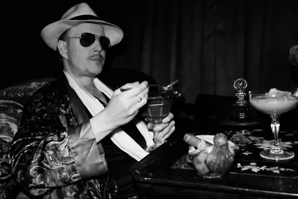 Un hombre con gafas de sol, sombrero de vaquero y una chaqueta estampada está sentado ante una mesa sosteniendo un vaso, con cócteles y objetos decorativos a su alrededor en blanco y negro.