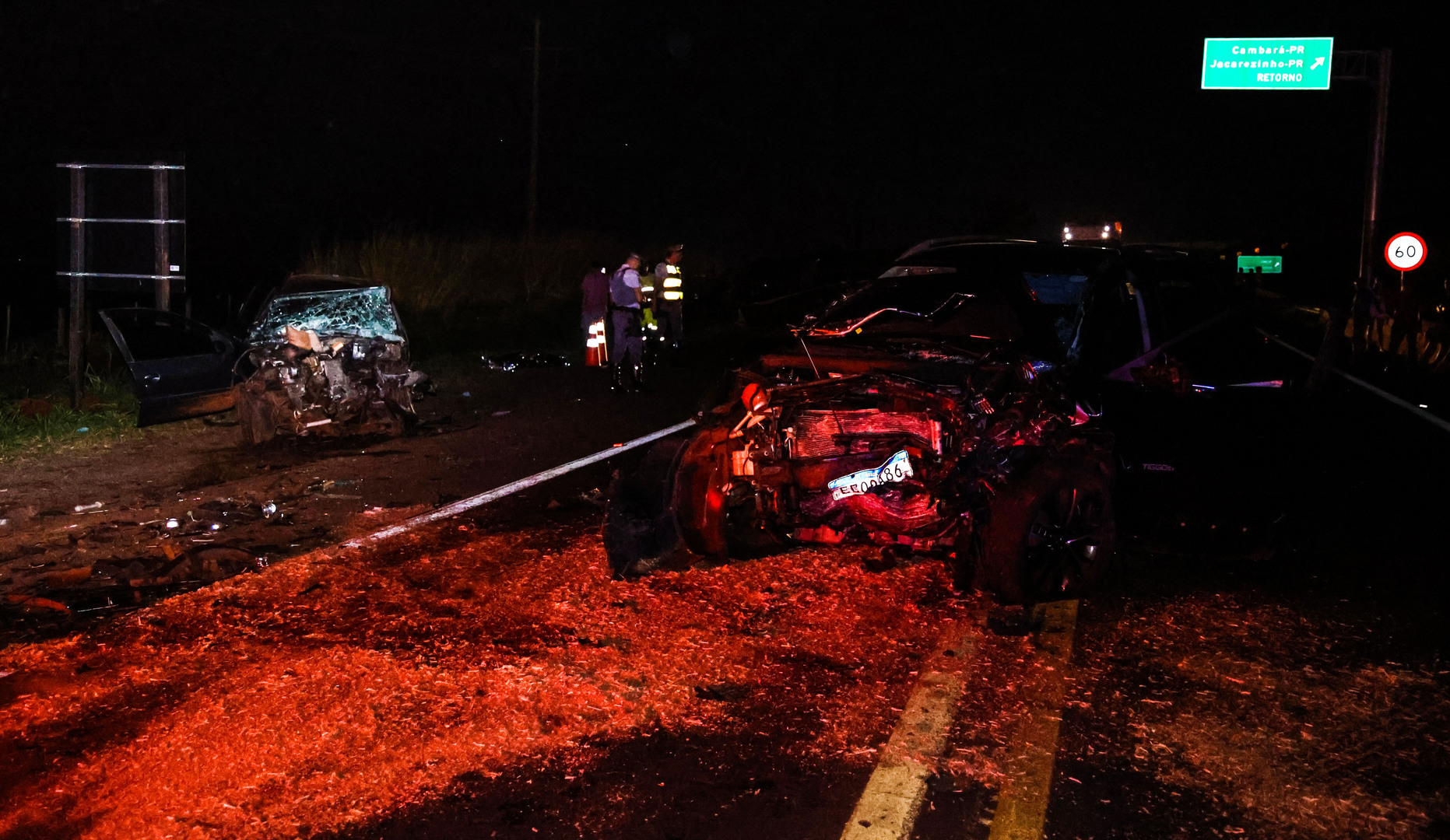 Two severely damaged vehicles at night on a highway, illuminated by red emergency lights, with debris scattered across the road and emergency responders in the background.