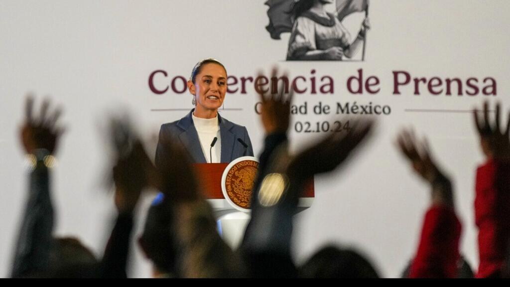 Una mujer en blazer azul habla en un podio con el escudo presidencial durante una conferencia de prensa en la Ciudad de México, con las manos de asistentes levantadas en primer plano.