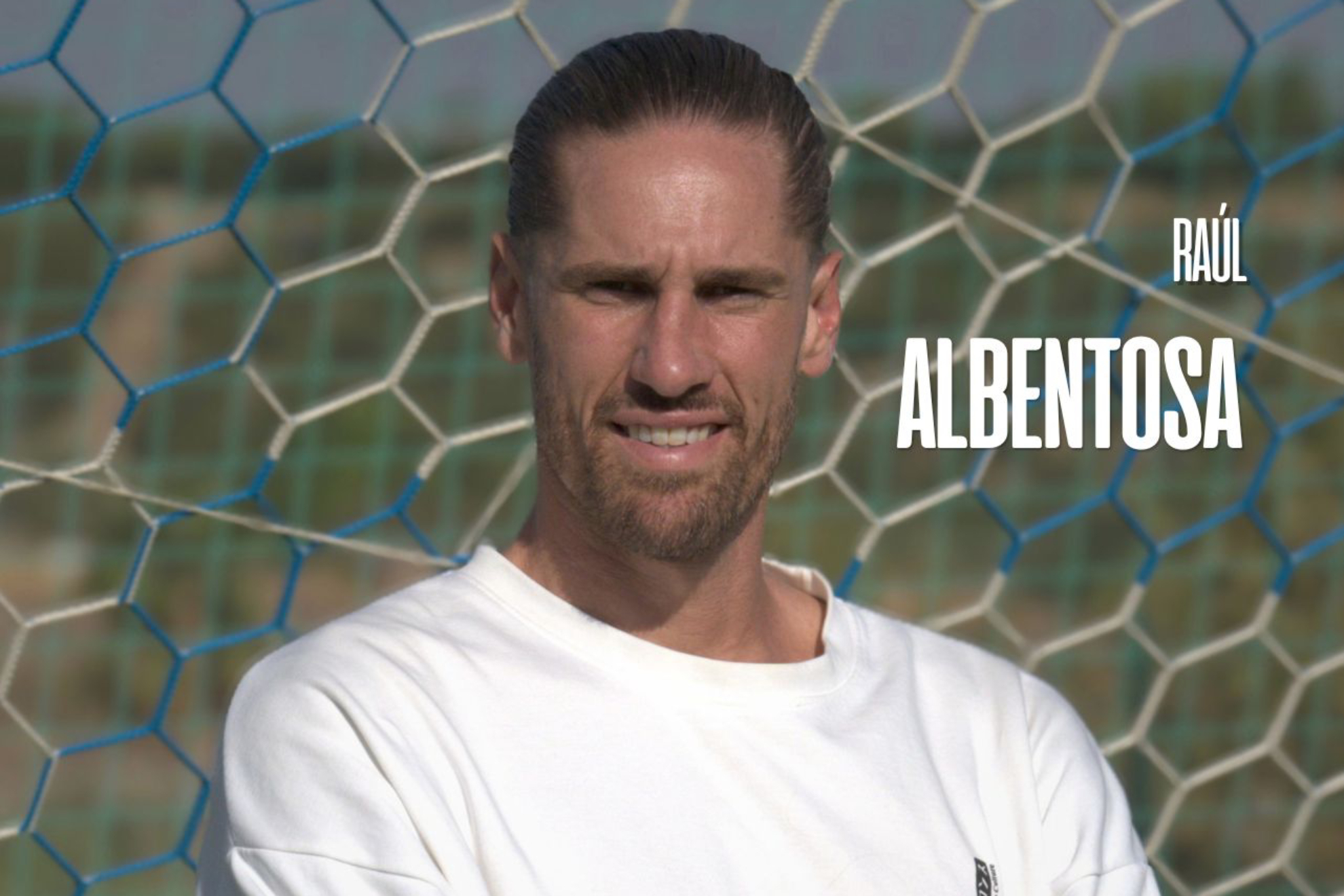 A man wearing a white t-shirt stands in front of a soccer goal net with blurred green grass in the background.