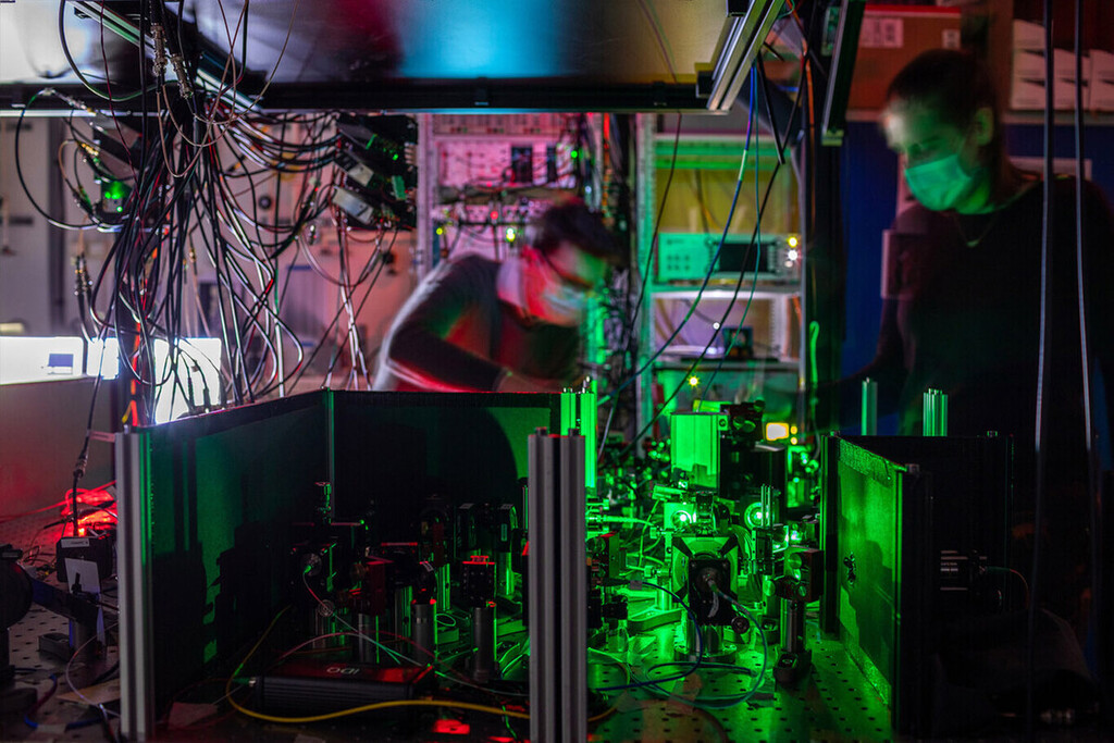 Un laboratorio de física cuántica con equipos de investigación iluminados en verde, cables enredados en la parte superior, monitores mostrando datos, y personal trabajando en el fondo.