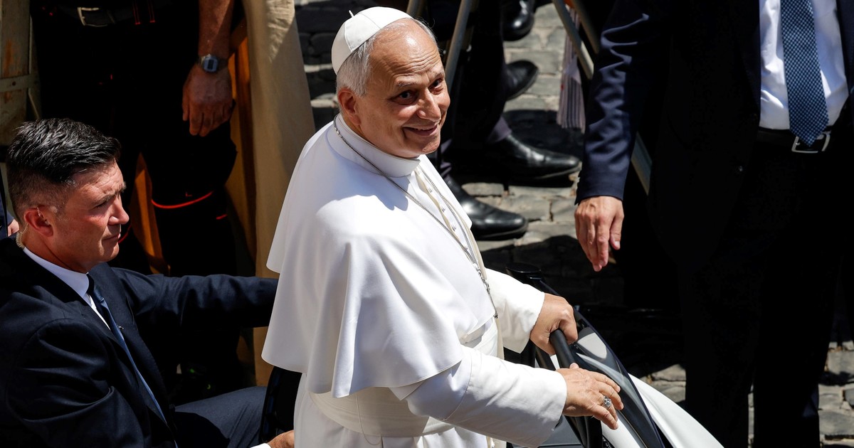 An elderly man wearing white vestments and a zucchet gets into a vehicle while surrounded by security personnel dressed in dark clothing.
