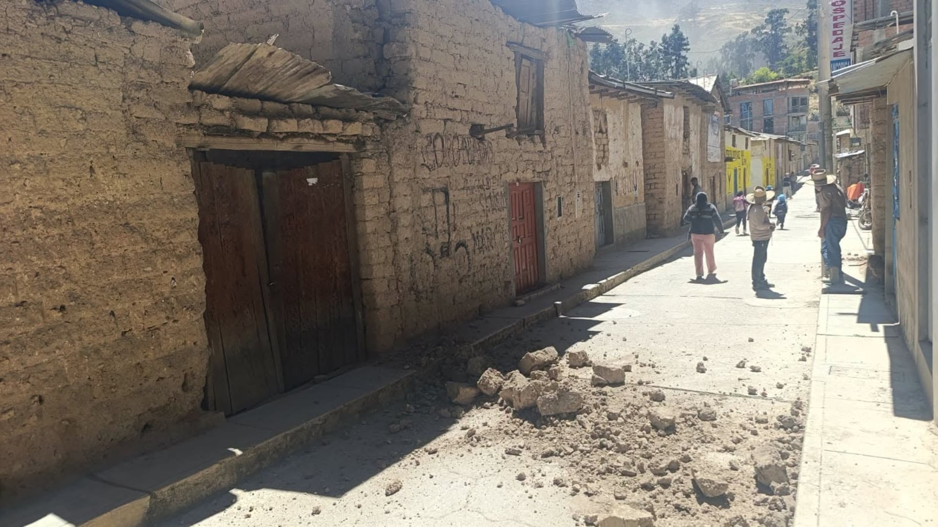 Una calle de un pueblo andino con casas de adobe dañadas, escombros dispersos en el suelo y personas caminando entre los edificios afectados.