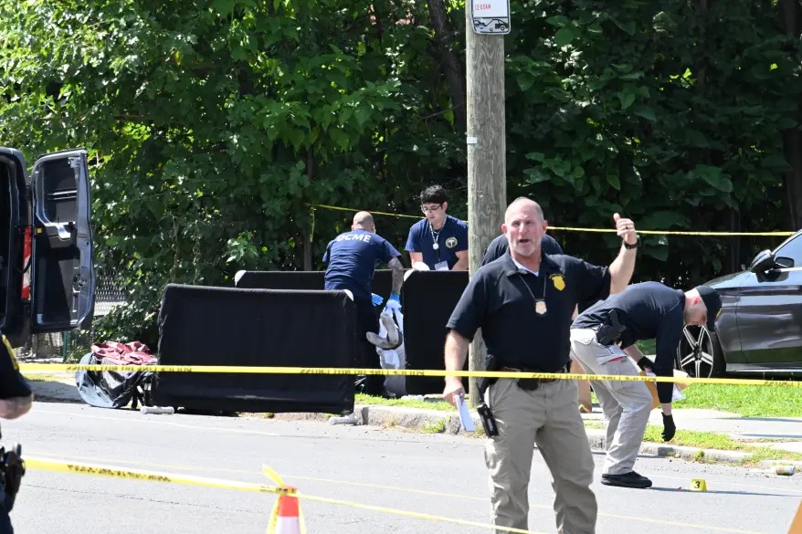Police officers and investigators work at a crime scene marked with yellow tape, with a black container and emergency vehicles visible on a tree-lined residential street.