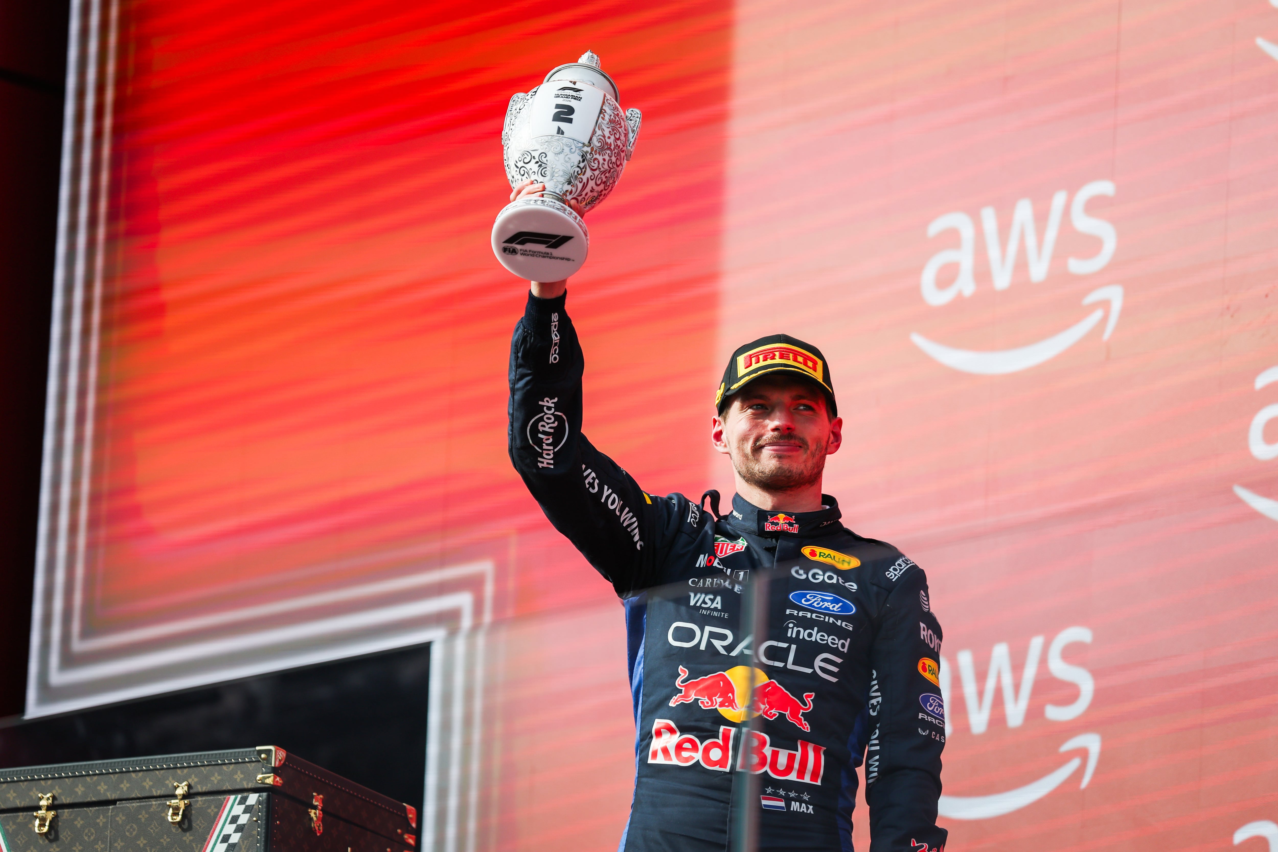 A Formula 1 driver in a Red Bull racing suit holds up a trophy while standing in front of a red screen displaying the AWS logo.