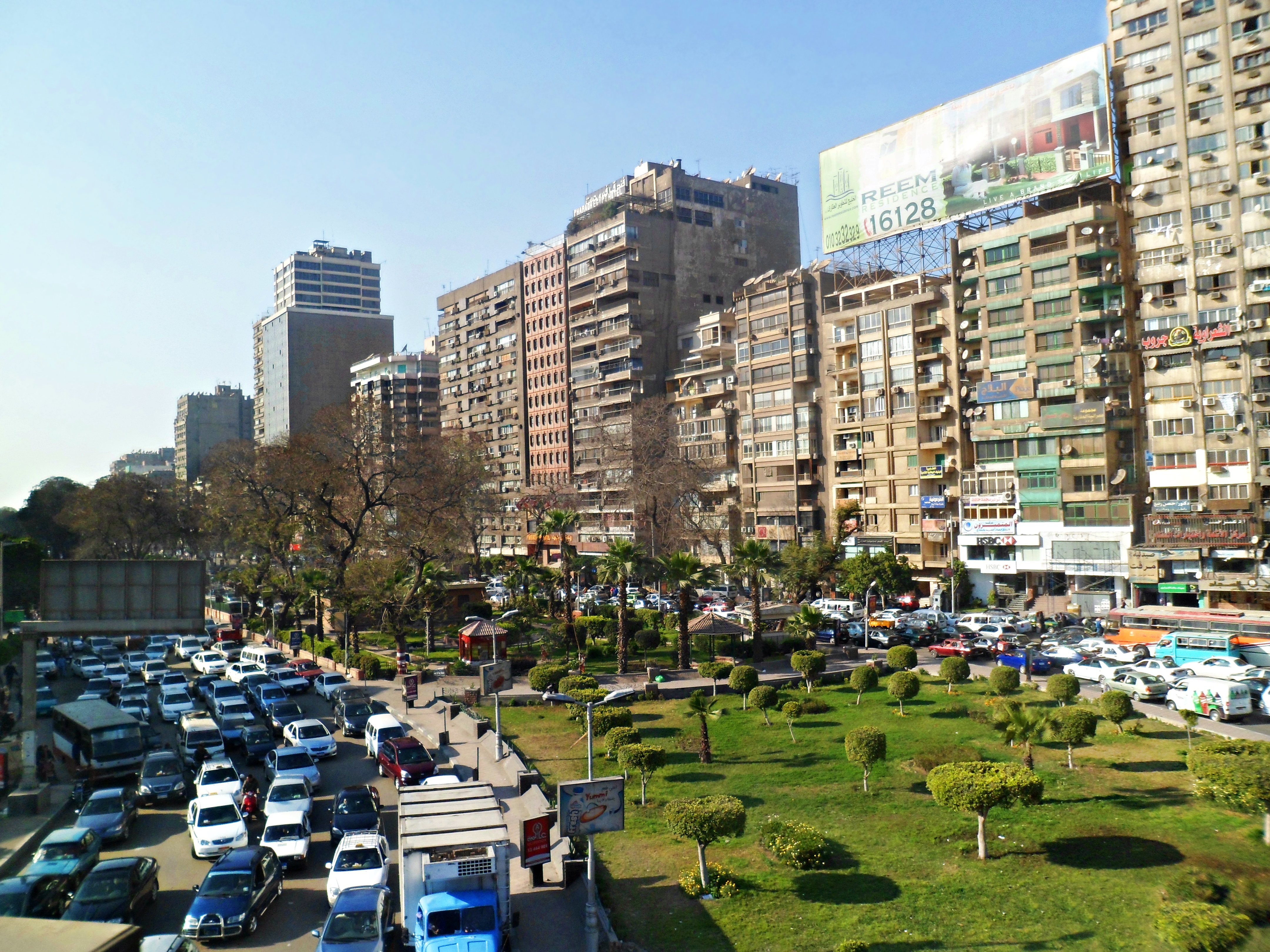 A busy urban plaza in Cairo with dense high-rise residential apartment buildings surrounding a tree-lined central square filled with parked cars and street-level shops.