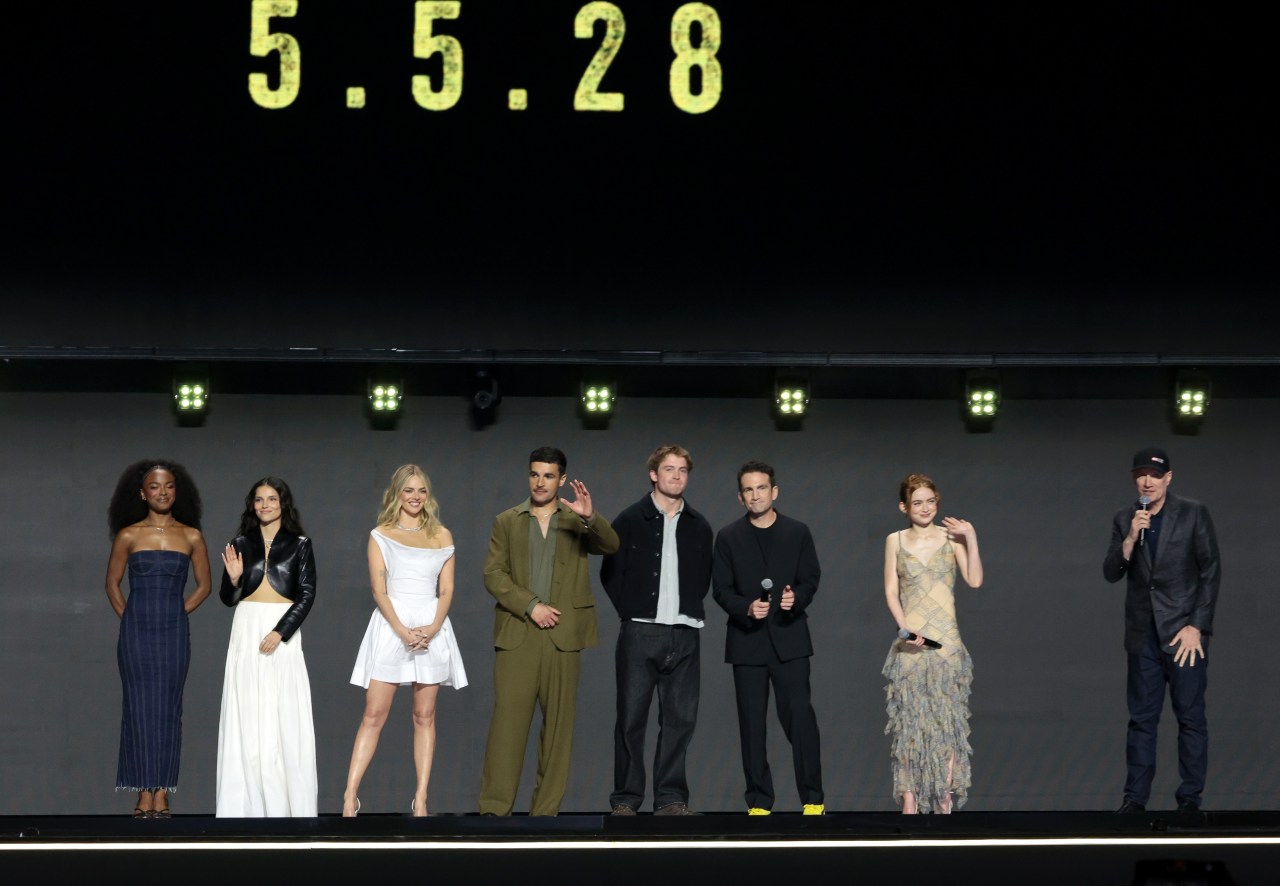 Nine cast members stand in a line on a stage with studio lighting, with a date "5.5.28" displayed on a screen above them.